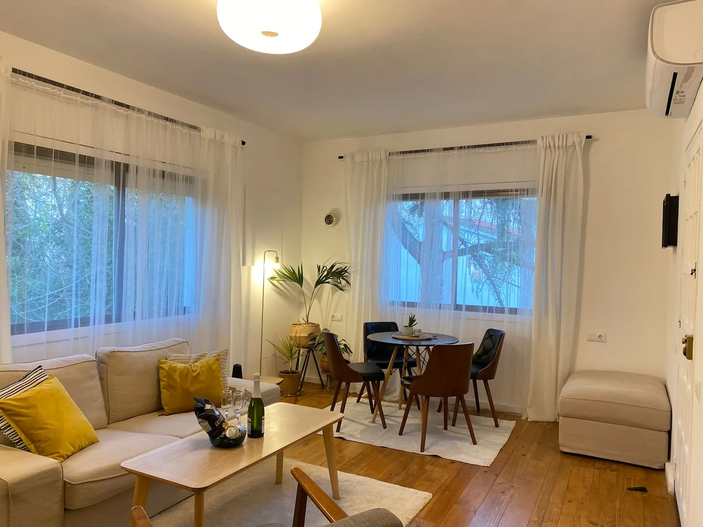 Living room with a beige sofa, round dining table and sheer curtains over warm wood floors