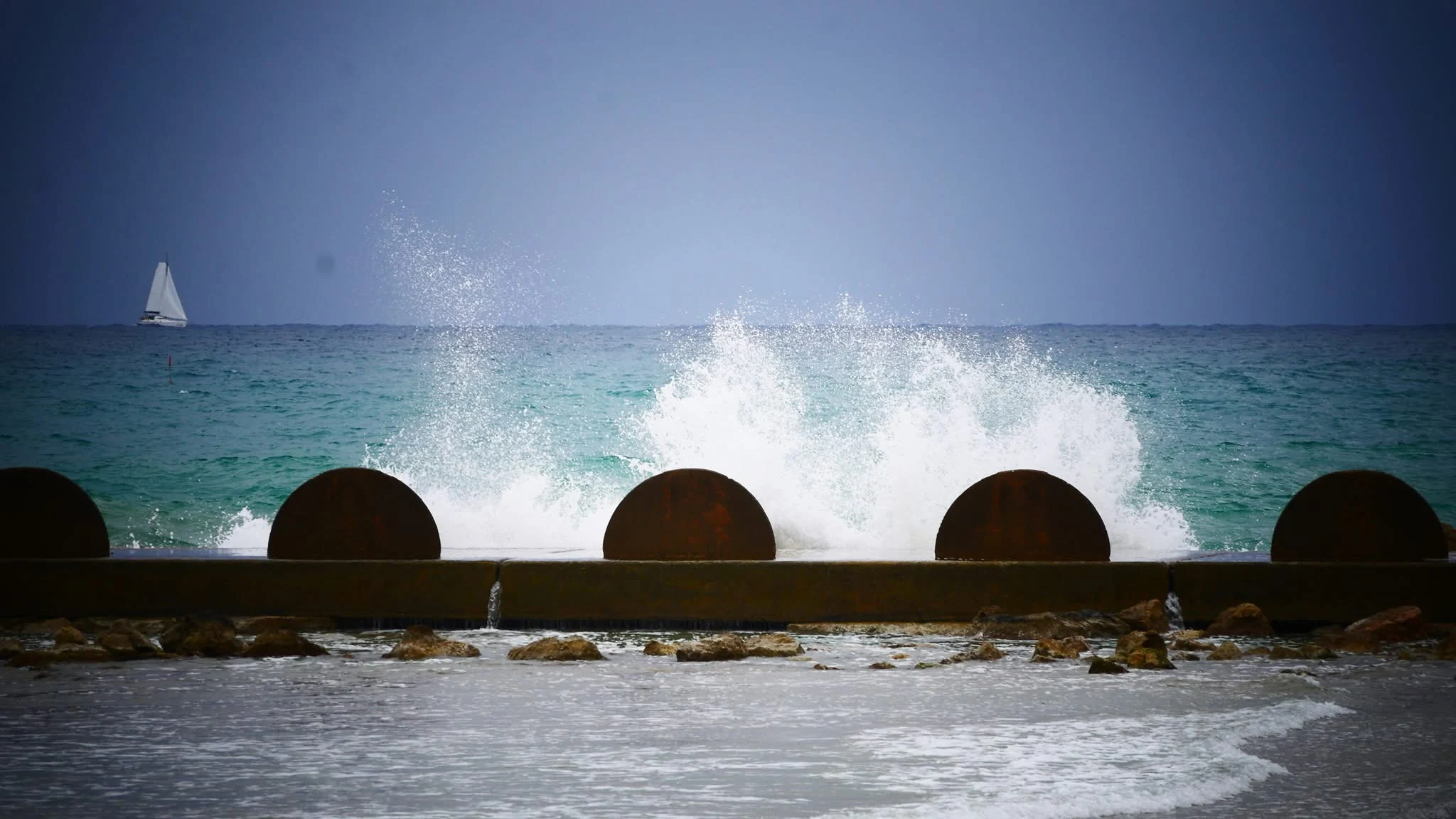 Waves breaking over the Cunit breakwater with a sailboat on the turquoise sea