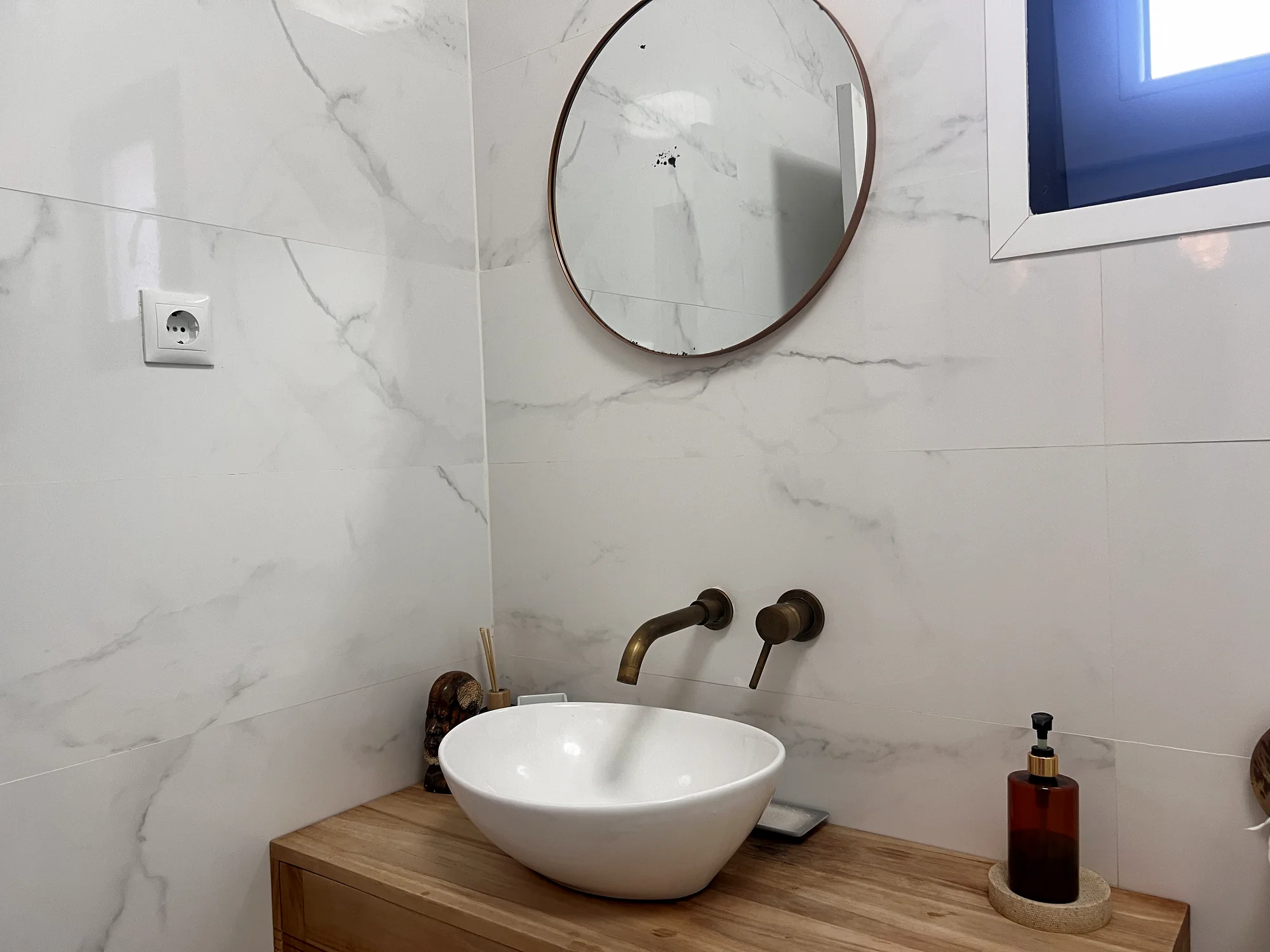 Bathroom vanity with a round mirror, wood counter, vessel basin and brushed-brass wall taps against marble tiling