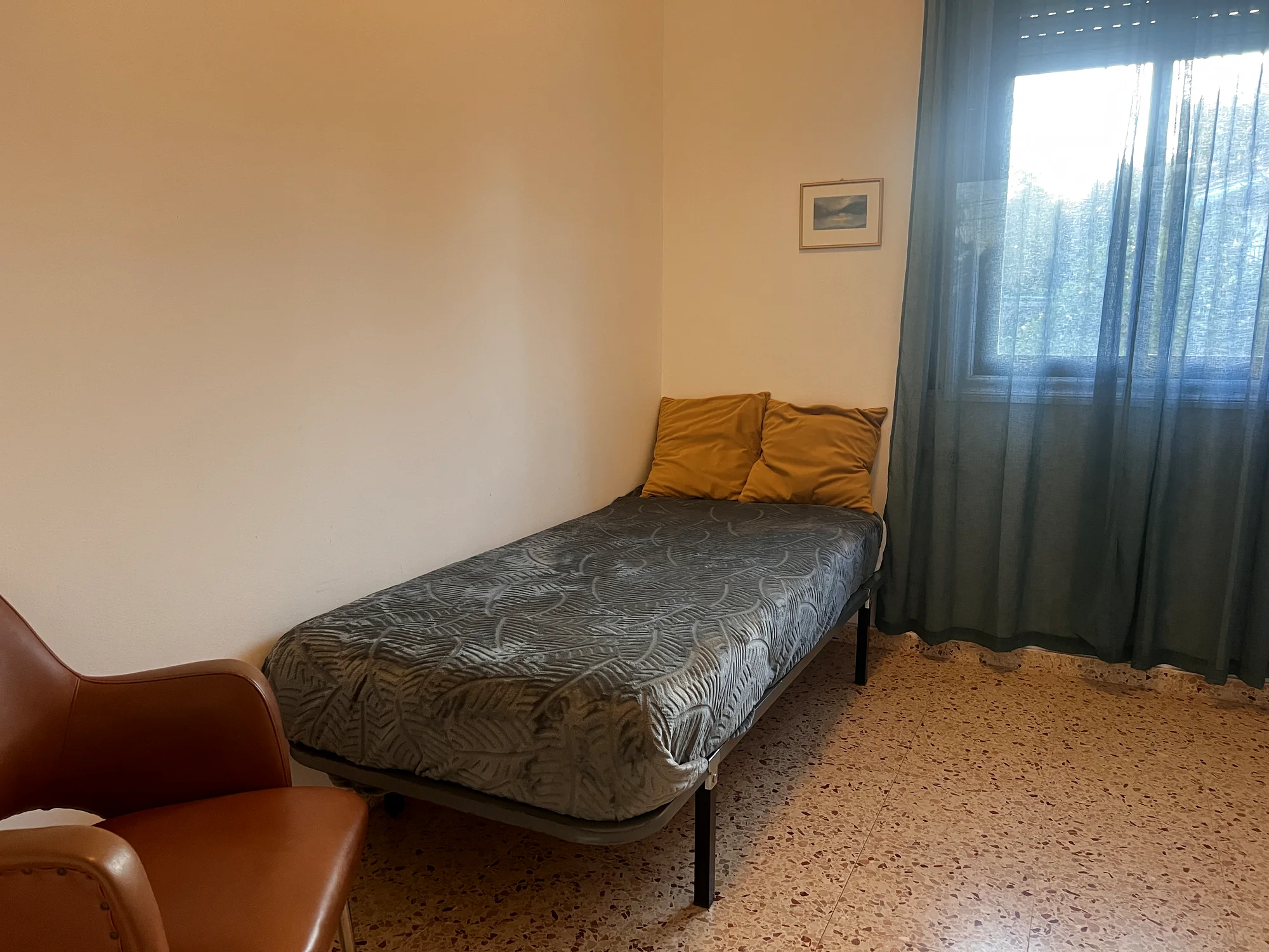 Single bedroom with a made-up bed, mustard pillows, a leather armchair and framed art by the curtained window