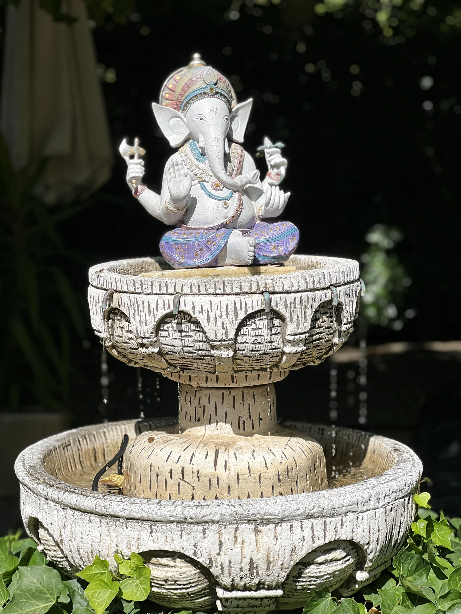 Two-tier stone Ganesha fountain framed by dark green foliage in the garden